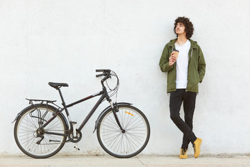 Horizontal shot of male cyclist takes break during bike ride, drinks takeaway coffee, dressed in fashionable clothes, thinks about something, shows his new modern bicycle, isolated on white background