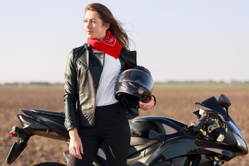 Fototapeta premium Relaxed carefree female biker looks thoughtfully aside, carries helmet, dressed in black leather jacket, stands near motorbike, has stop in road, focused into distance. People, ride and morocycling