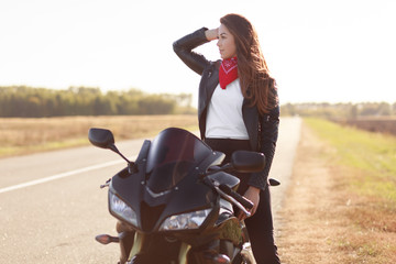 Pensive female driver in stylish clothes, poses on fast motorbike, looks thoughtfully aside, being active biker, poses in countryside, thinks about new extreme destination. People, lifestyle concept