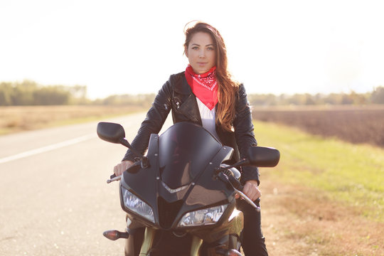 Outdoor shot of beautiful motorcyclist has long journey on her motorbike, wears red stylish bandana and leather jacket, likes extreme driving, poses on road, People, lifestyle and hobby concept
