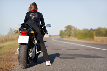 Obraz premium Back view of stylish biker on motorbike, covers long destination, rides in countryside in open road, enjoys time for relax, likes her transport, feels freedom and independence. Outdoor activity