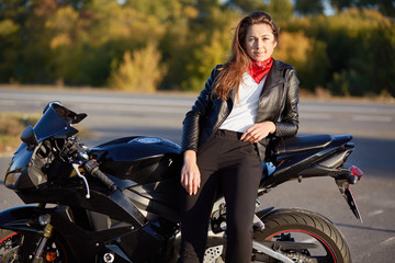 Fototapeta na wymiar Photo of self assured woman dressed in leather jacket, black trousers, leans on motorbike, has thoughtful expression, rests after long journey, has good rest, poses outdoor against blurred background
