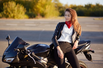 Fototapeta na wymiar Horizontal view of thoughtful biker stands near favourite motorbike, dressed in stylish outfit, has contemplative look down, breathes fresh air, likes extreme spot. People, lifestyle and active rest