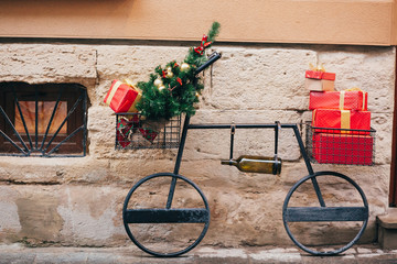 Fototapeta na wymiar Creative bicycle with christmas tree,red presents, wine bottle in european city street. Stylish christmas street decor, Festive decorations and illumination. Winter holidays. Wine shop.