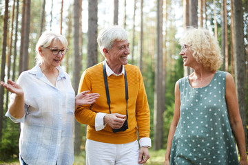 Senior man with photocamera and two happy women discussing where to go during chill in the forest