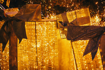 Golden gift boxes with lights under christmas tree in evening european city center. Big presents with bows and illumination on background of lights. Festive decor in winter holidays