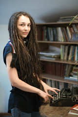 Fototapeta na wymiar girl with dreadlocks in a scarf standing in the library and typing on a typewriter