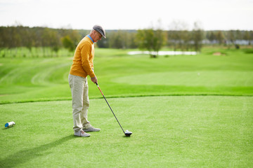 Senior casual man standing on green lawn and keeping golf club close to ball on grass