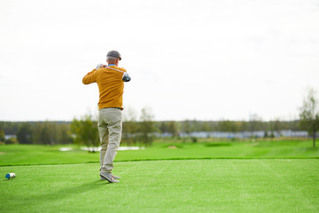 Back view of senior man in cap, yellow pullover and beige pants hit ball with golf club on green...