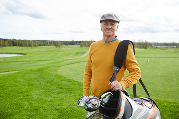 Aged successful golf player with bunch of clubs in sportive bag on his shoulder looking at you