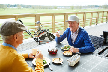 Two mature golf players sitting by table in front of one another, having salad and tea and...