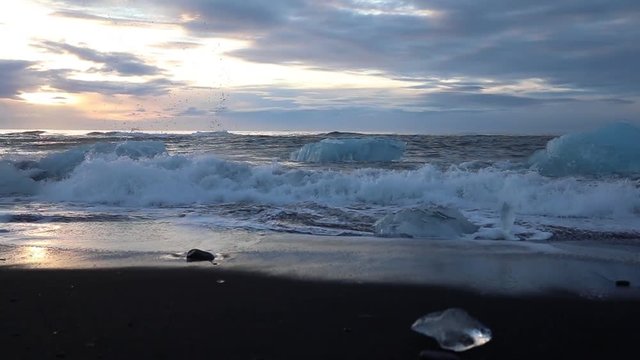 Icebergs and pieces of blue and white ice are rolling on the waves. Waves breaking on the ice. Surf on the beach at sunrise. Iceland