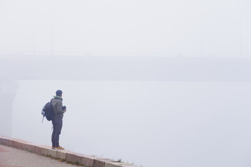 Man photographer looking at bridge holding camera in his hands on foggy morning. Rear view. Copy space.