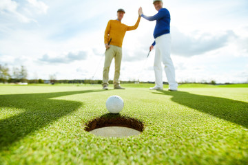 Two active men making high five while standing on play field and looking at golf ball by hole