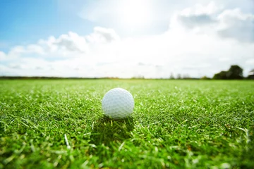 Wall murals Golf White golf ball laying on green grass of play field on sunny day with cloudy sky above