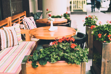 Cozy outdoor summer space in a cafe. Comfortable chairs and classic wooden round tables. Red flowers in the flowerpots. Colorful atmospheric background
