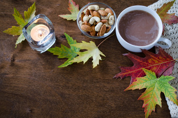 A cup of hot cocoa with knitted blanket, nuts, candle and autumn leaves on a rustic wooden table.