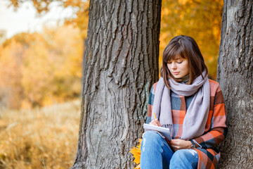 University Park, study concept. Portrait of young woman in fall forest . Writes in a notebook. Brunette woman in autumn park with fashionable plaid coat and scarf.