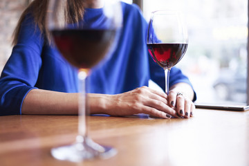 Woman is drinking red wine and having rest in cafe near window, close-up of woman’s hands holding glass on table.