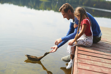 Young man and his daughter feeding duck while sitting on pontoon by waterside © pressmaster