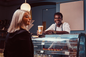 Smiling African American barista in uniform giving a cup of coffee to his client at the trendy coffee shop.