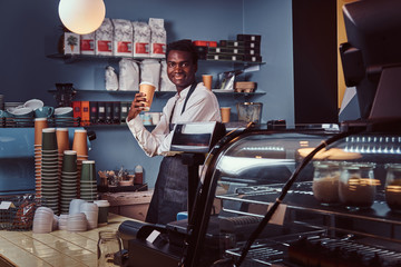 Handsome African American barista in uniform holds cup of coffee while standing in his coffee shop.