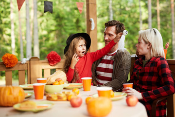 Little girl in halloween attire frightening her mom with toy ghost during family dinner on terrace