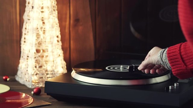A young woman in a red knitted winter pullover puts a black vinyl record on a turntable, a stylus a needle and listens to music against the background of a glowing Christmas tree and a cozy New Year's