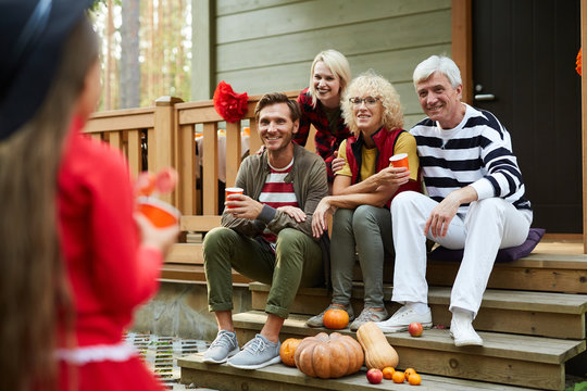 Two couples sitting on terrace of country house and looking at litle girl in halloween attire asking for treats