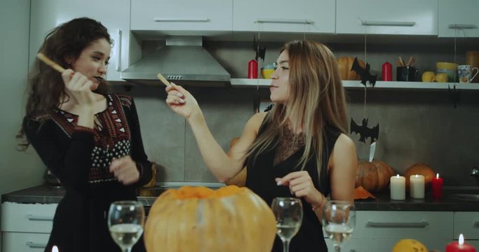 Playing two ladies in front of the camera at Halloween party at home , holding something in hand , they are smiling.