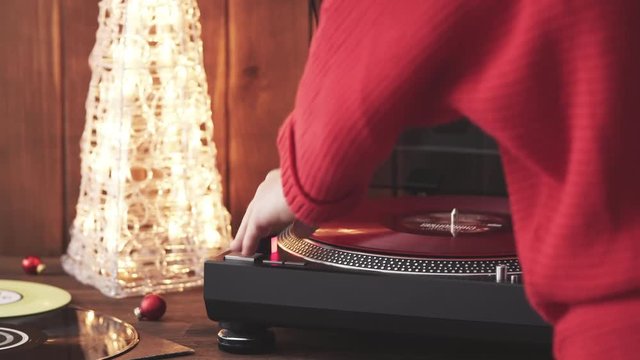 A young woman in a red knitted winter pullover puts a red vinyl record on a turntable, a stylus a needle and listens to music against the background of a glowing Christmas tree and a cozy New Year's