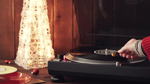 A young woman in a red knitted winter pullover puts a black vinyl record on a turntable, a stylus a needle and listens to music against the background of a glowing Christmas tree and a cozy New Year's