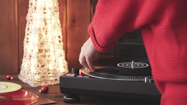 A young woman in a red knitted winter pullover puts a black vinyl record on a turntable, a stylus a needle and listens to music against the background of a glowing Christmas tree and a cozy New Year's