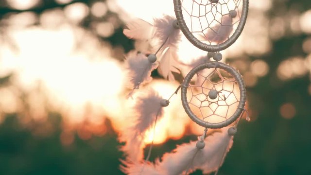Slow motion amulet dreams catcher waving in a light wind against the backdrop of a bright sunset and a bokeh of city lights. White light feathers rise in the wind. The disc of the setting sun. Ethnic