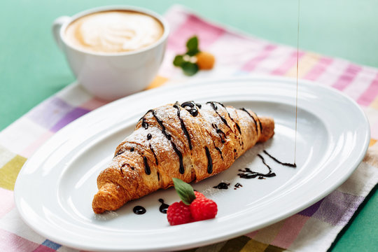 Fresh homemade croissants and cappuccino on cloth napkin decorated with raspberry and chocolate topping. Side view with copy space on green background