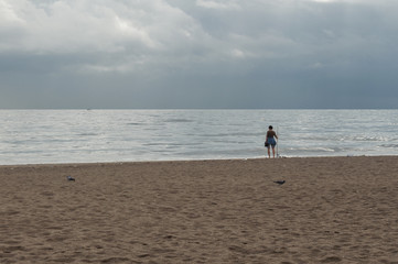 Lonely woman standing with a stick looking the sea while a storm is approaching. Cloudy day in a beach with birds in the sand. Stormy landscape.