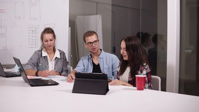 Young man in glasses and two women sitting at the office table in creative workplace. Brunette woman pointing on whitesheet board, sharing her vision. Man got up and continue to explaine near the
