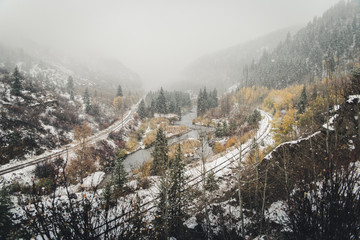 Landscape view of a railroad track and river surrounded by fall foliage during a snow storm. 