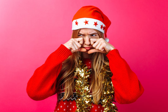 Upset girl in Santa's hat and with tinsel on her neck, rubs her eyes and wants to cry with resentment on a red background.