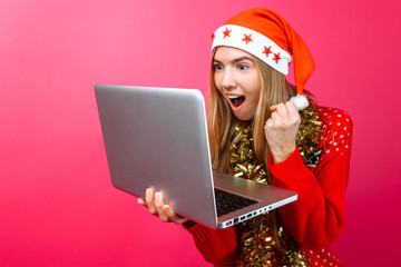 Happy girl in red sweater and hat of Santa looking at a laptop excited about the good news , the shocked girl shows gesture success victory, on a red background.