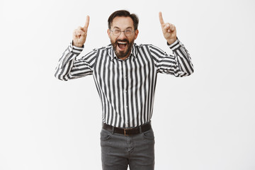 Naklejka premium Guy yelling from happiness becoming number one in company. Pleased and triumphing good-looking emotive adult man in formal outfit and glasses, shouting at camera and pointing up with index fingers