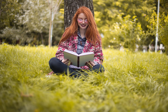 Redhead cheerful woman with freckles and dark lipstick wearing sunglasses reading book outdoors in park sit under big tree on grass with flowers on the background.