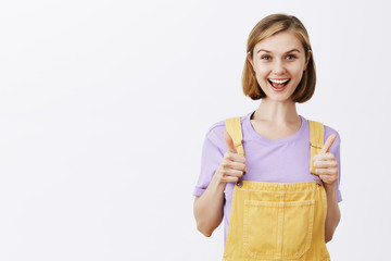 Yeah I support your idea. Attractive young and stylish happy female student in yellow overalls, showing thumbs up and smiling joyfully while liking great suggestion, cheering and supporting friend