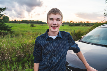 Happy young man leaning against his car next to an open field at sunset.