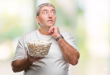 Handsome senior man eating popcorn over isolated background serious face thinking about question, very confused idea