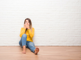 Young adult woman sitting on the floor over white brick wall shocked covering mouth with hands for mistake. Secret concept.