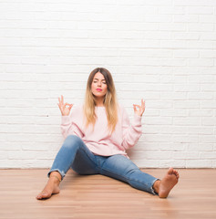 Young adult woman sitting on the floor over white brick wall relax and smiling with eyes closed doing meditation gesture with fingers. Yoga concept.