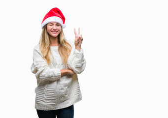 Young beautiful blonde woman wearing christmas hat over isolated background smiling with happy face winking at the camera doing victory sign. Number two.