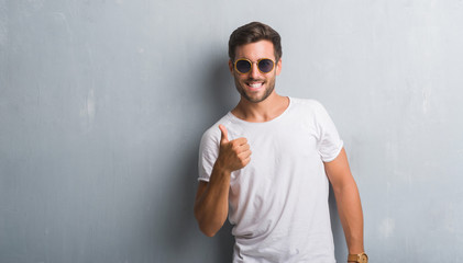 Handsome young man over grey grunge wall wearing sunglasses doing happy thumbs up gesture with...