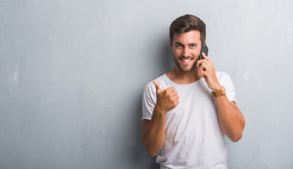 Handsome young man over grey grunge wall speaking on the phone happy with big smile doing ok sign, thumb up with fingers, excellent sign